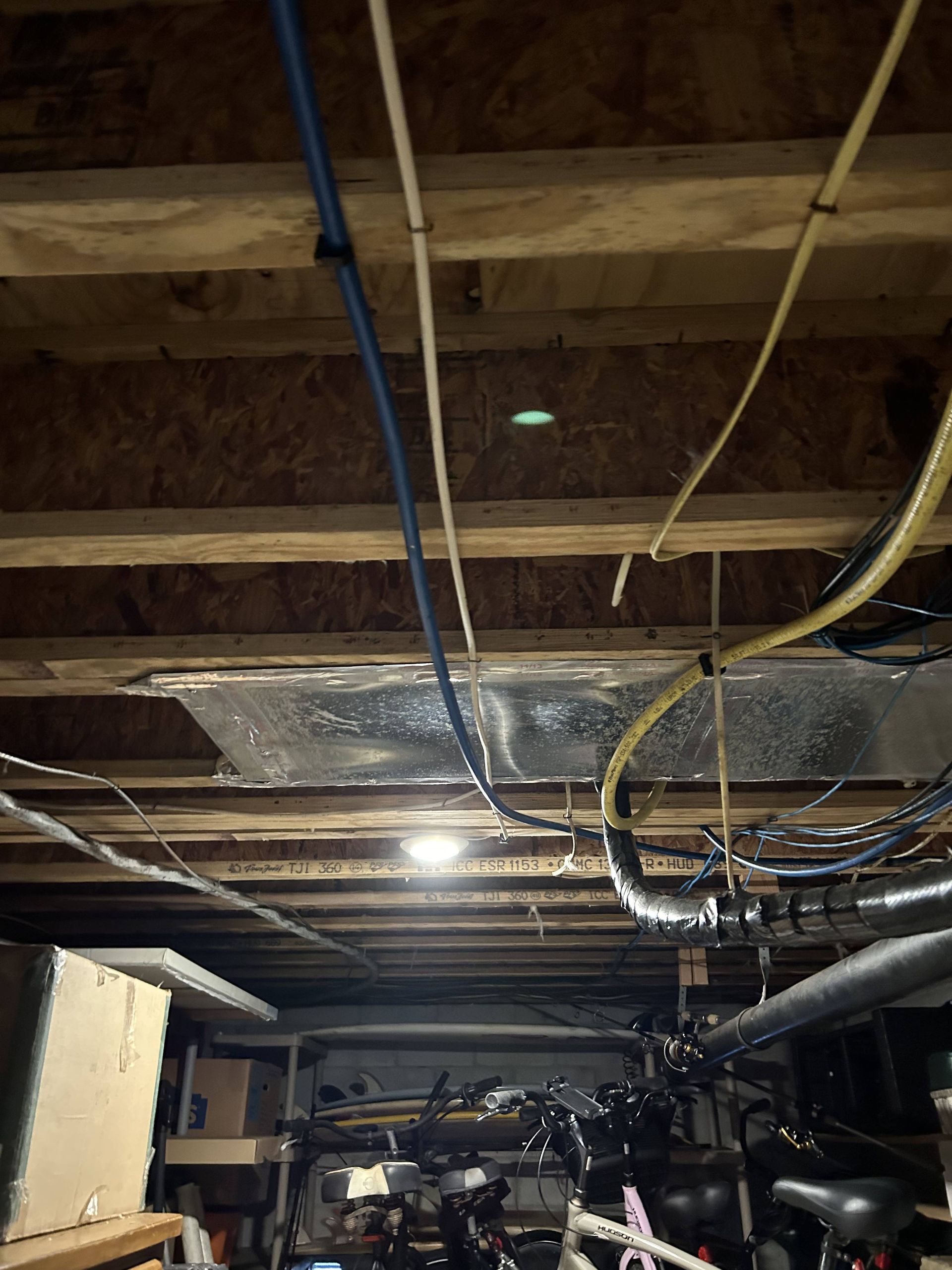 Basement ceiling with exposed wooden beams, hanging cables, and metal ductwork over stored equipment