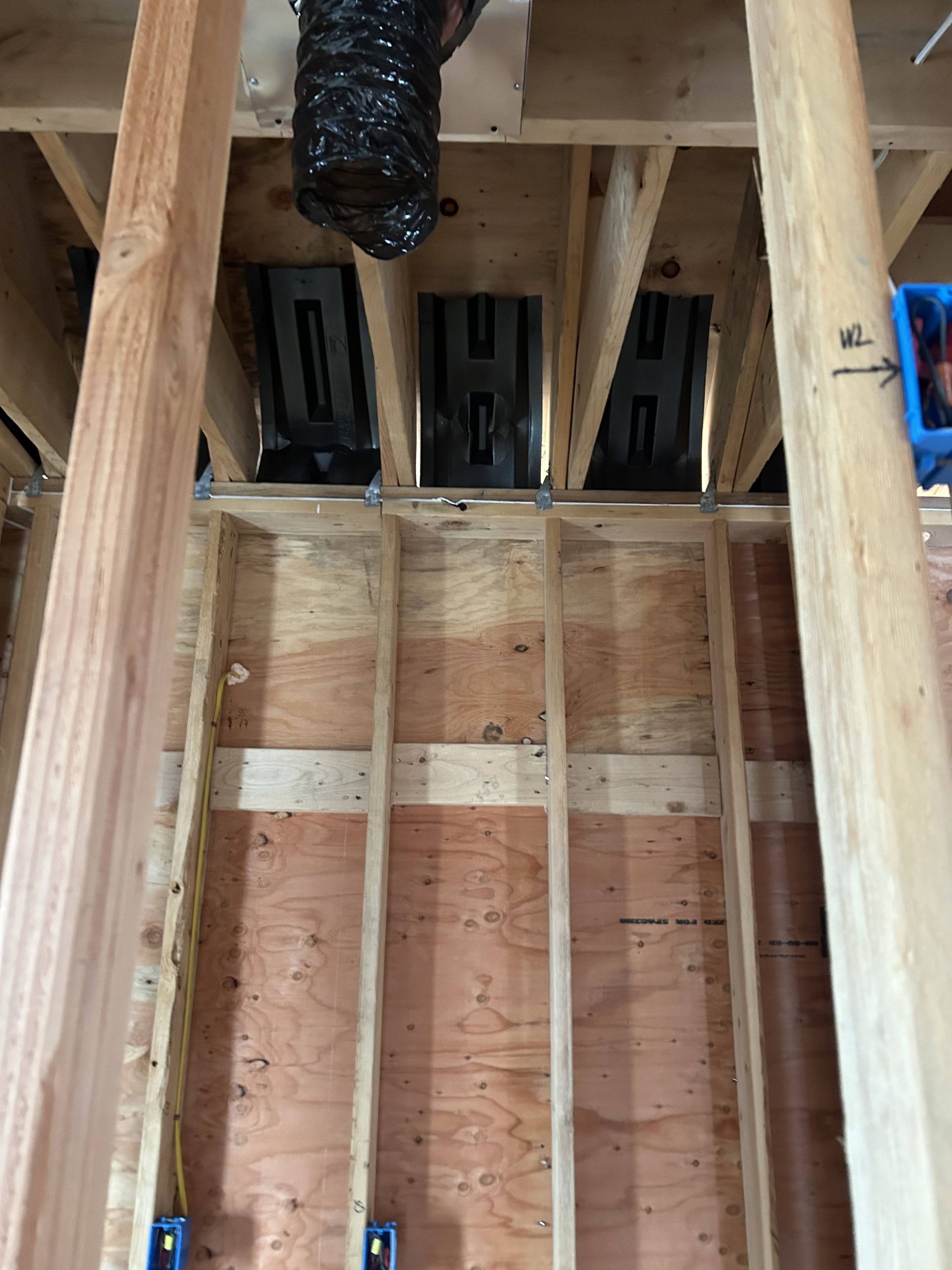 Exposed wooden wall framing with plywood sheathing and black ductwork in an unfinished room