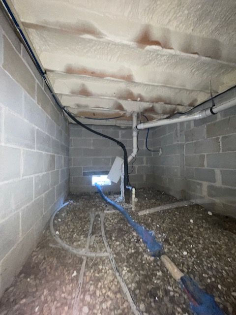 Crawlspace beneath a house with cinder block walls, exposed insulation, and plumbing pipes along the dirt floor.