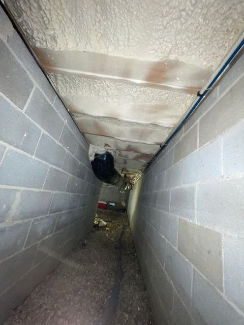 Narrow cinder block crawlspace with a low, damp ceiling and exposed pipes, viewed down a gravel floor.