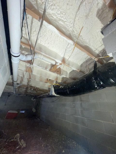 Crawlspace under a house with exposed pipes, insulation, and a black duct along a cinder block wall