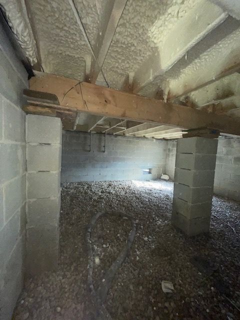 Crawl space with cinder block supports, exposed insulation, and gravel floor under a wooden beam.