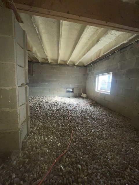 Unfinished basement with exposed ceiling beams, cinder block walls, gravel floor, and a small window