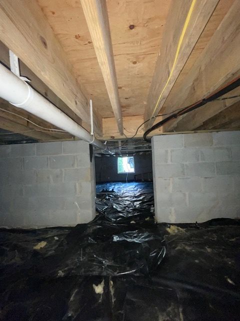 Crawl space with exposed floor joists, cinder block walls, black vapor barrier, and a white pipe overhead