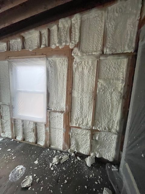 Foam insulation sprayed between wall studs around a small window, with debris on the floor.