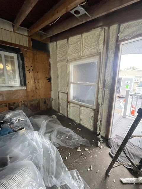 Damaged room with exposed insulation, plastic-covered debris, and a broken window by a doorway