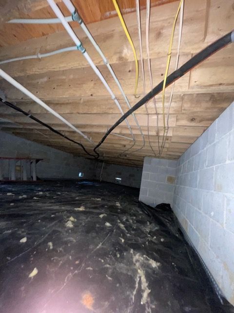 Crawlspace with exposed wooden joists, hanging wires, black vapor barrier floor, and cinder block wall
