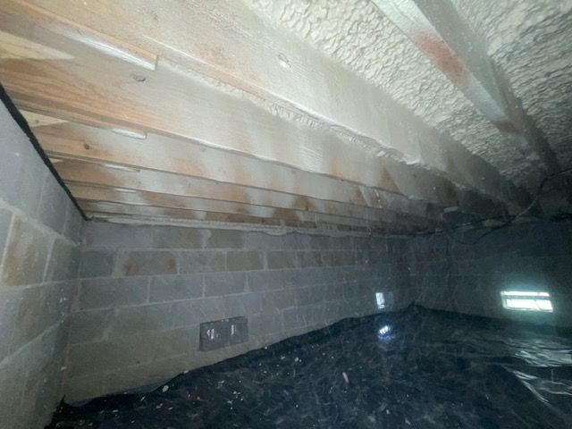 Insulated attic crawlspace with exposed wooden rafters, cinder block wall, and dark plastic-covered floor