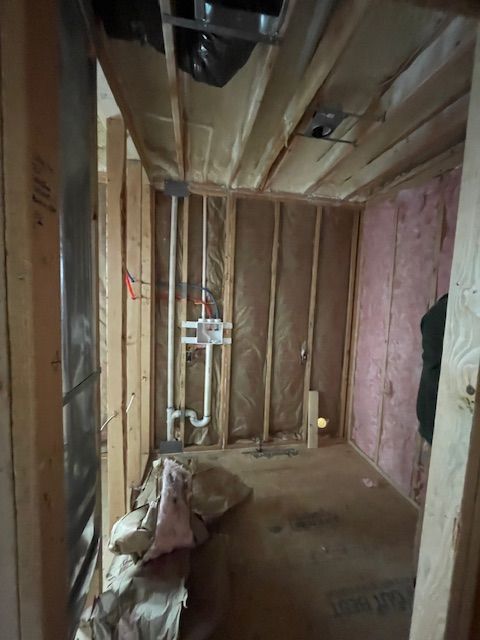 Unfinished interior wall framing with exposed pipes, pink insulation, and a bag of materials on the floor.