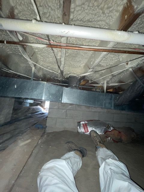 Looking down through joists into a crawl space, with two white pipes and a person’s legs visible at the bottom.