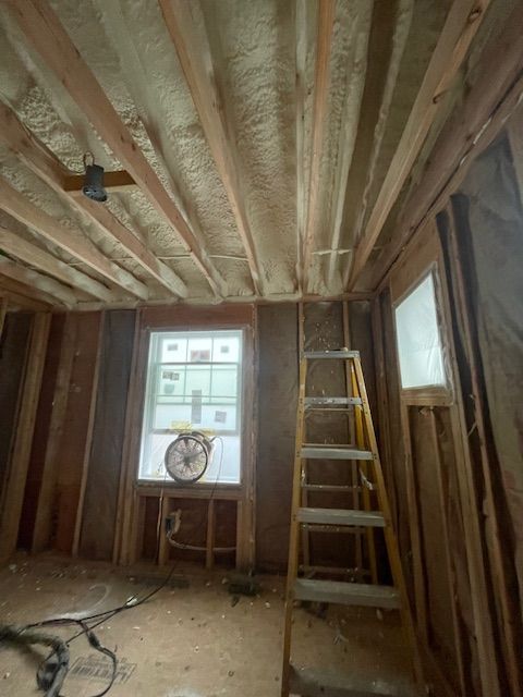 Interior of a room under renovation, with exposed insulation, a ladder, and a window with a fan.