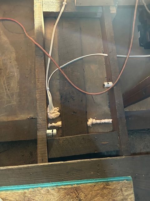 Exposed wiring and pipes inside a wooden wall cavity with red, white, and black cables.