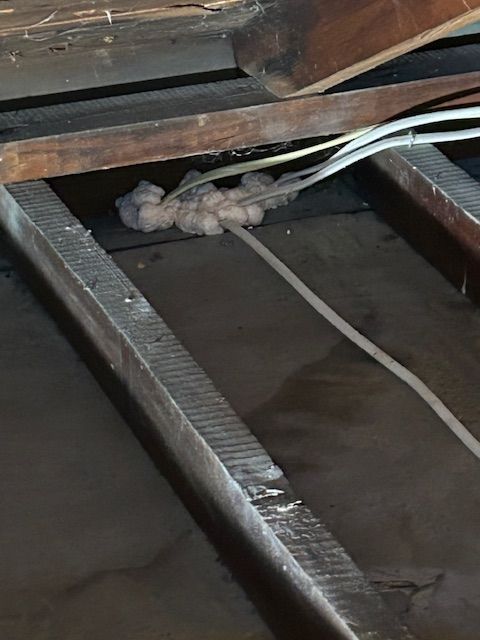 Exposed wooden beams and white cables in a dim crawl space with insulation stuffing.
