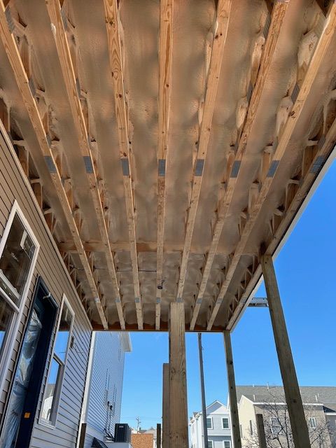 Wooden deck framing under construction beside a house, viewed from below against a blue sky.