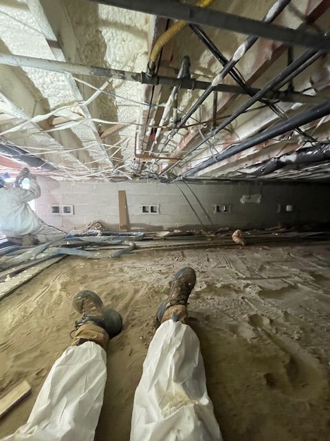 Person in white coveralls lies on a dusty crawlspace floor beneath exposed ductwork and pipes.