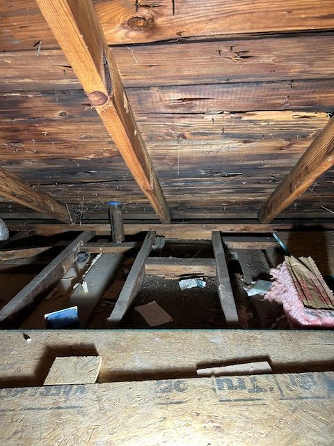 Exposed attic rafters and wooden planks with insulation and debris on the floor.