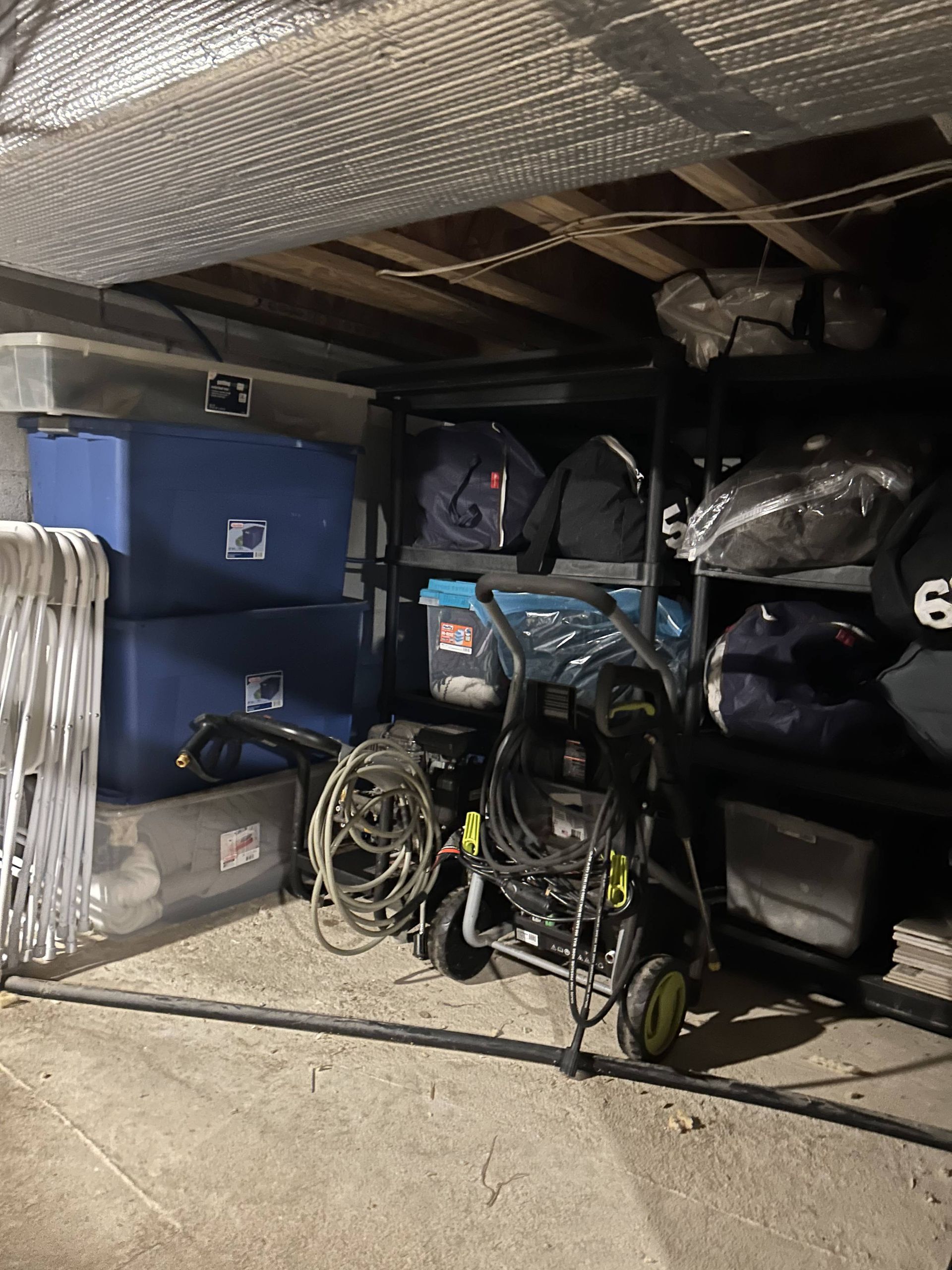 Cluttered storage space with blue bins, coiled cords, bags, and tools under a low insulated ceiling