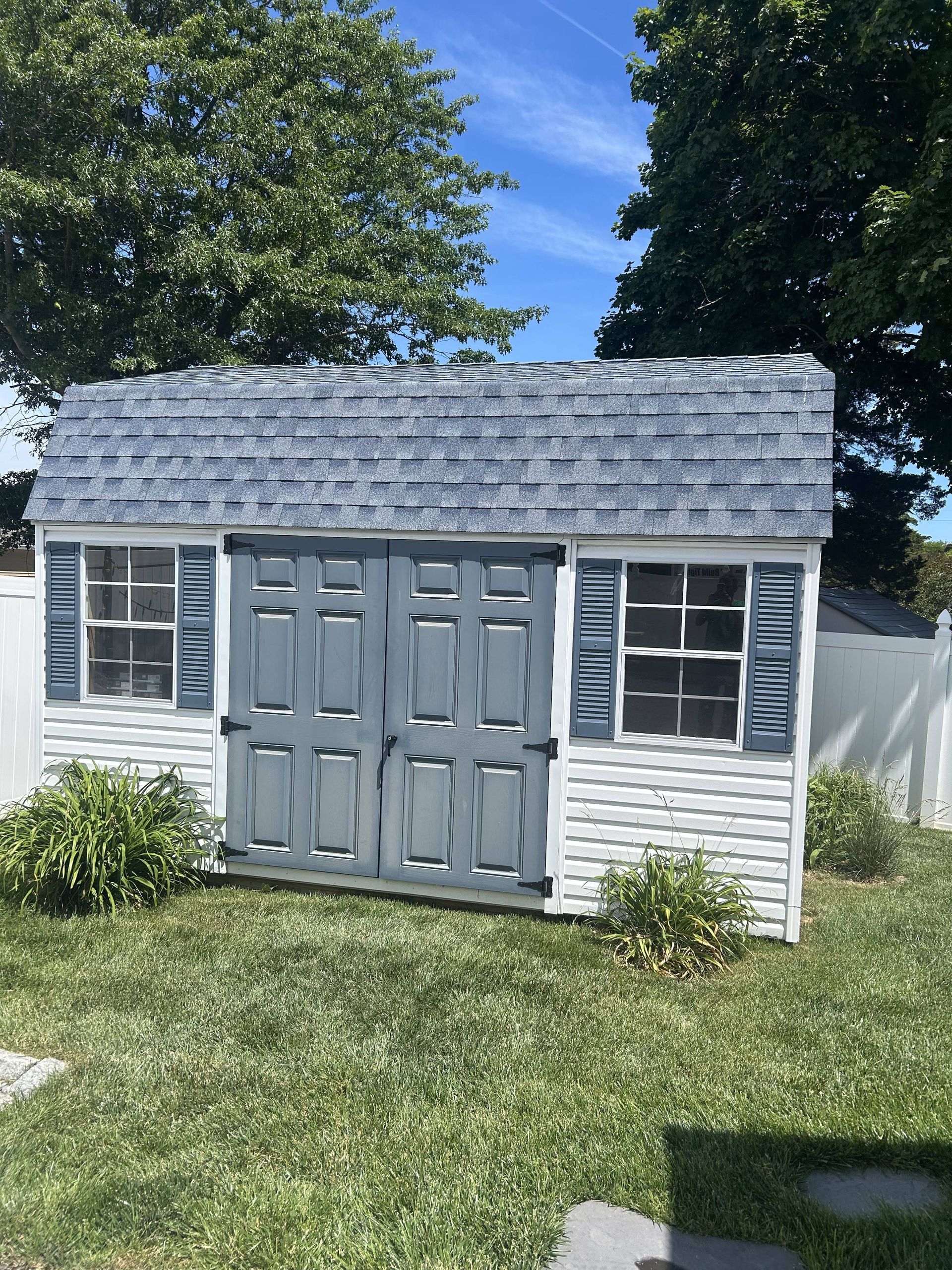 Small gray backyard shed with double doors, windows, and shingled roof in a grassy yard.