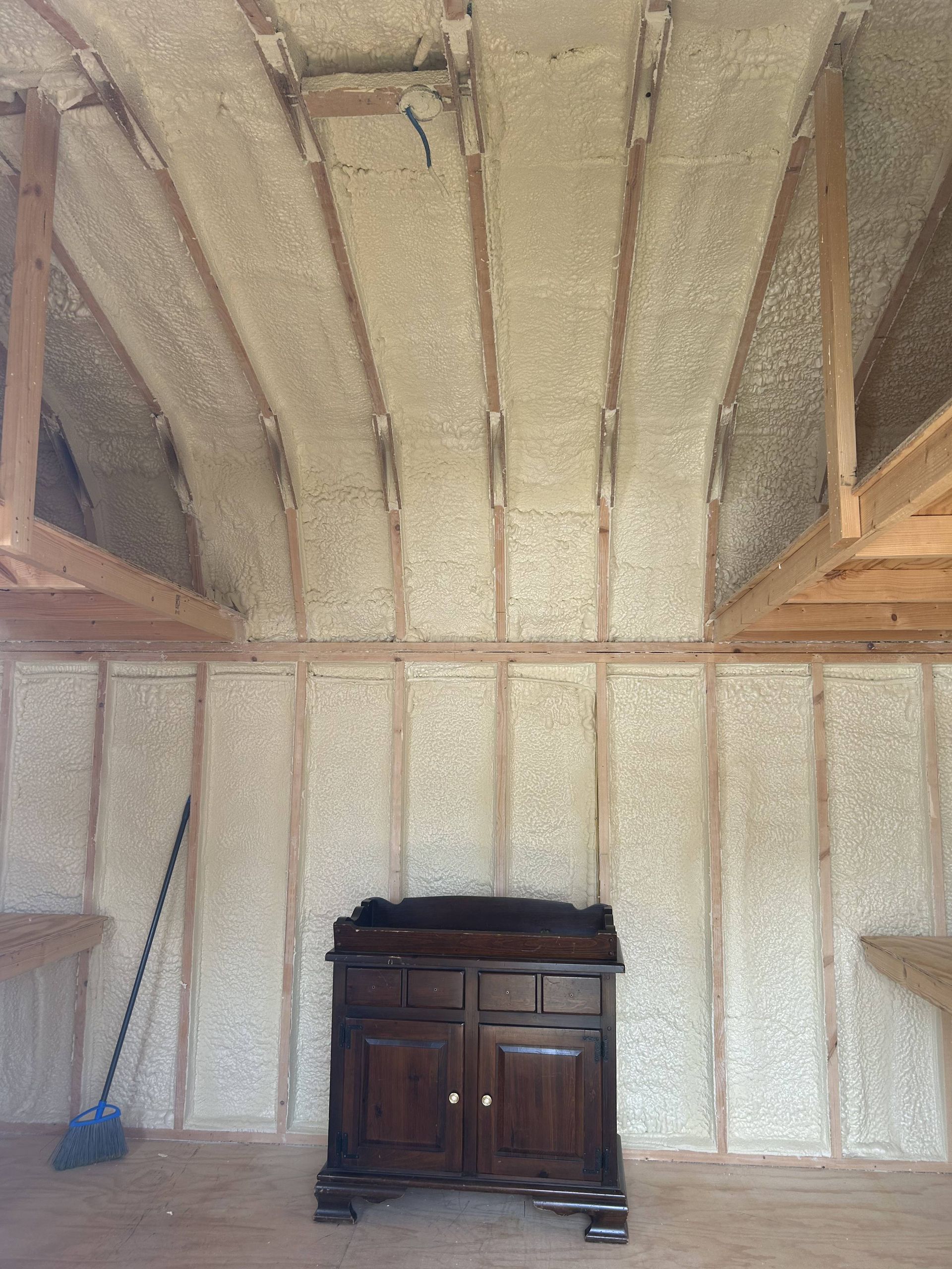 Unfinished room with exposed insulation, wooden beams, a small cabinet, and a blue broom leaning against the wall.