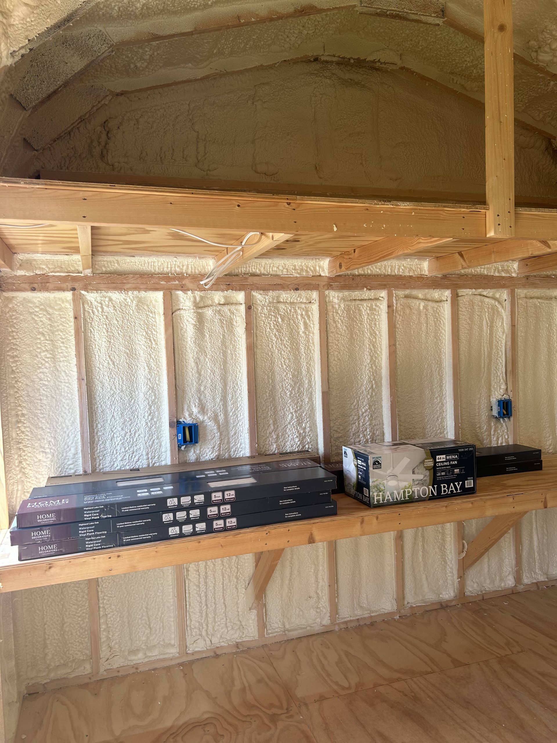 Interior under construction with spray foam insulation, wooden framing, and shelves holding tools and materials.