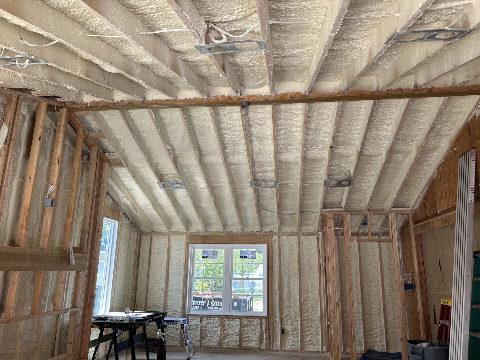 Interior of a room under construction, with spray foam insulation, exposed framing, and a window at the far end