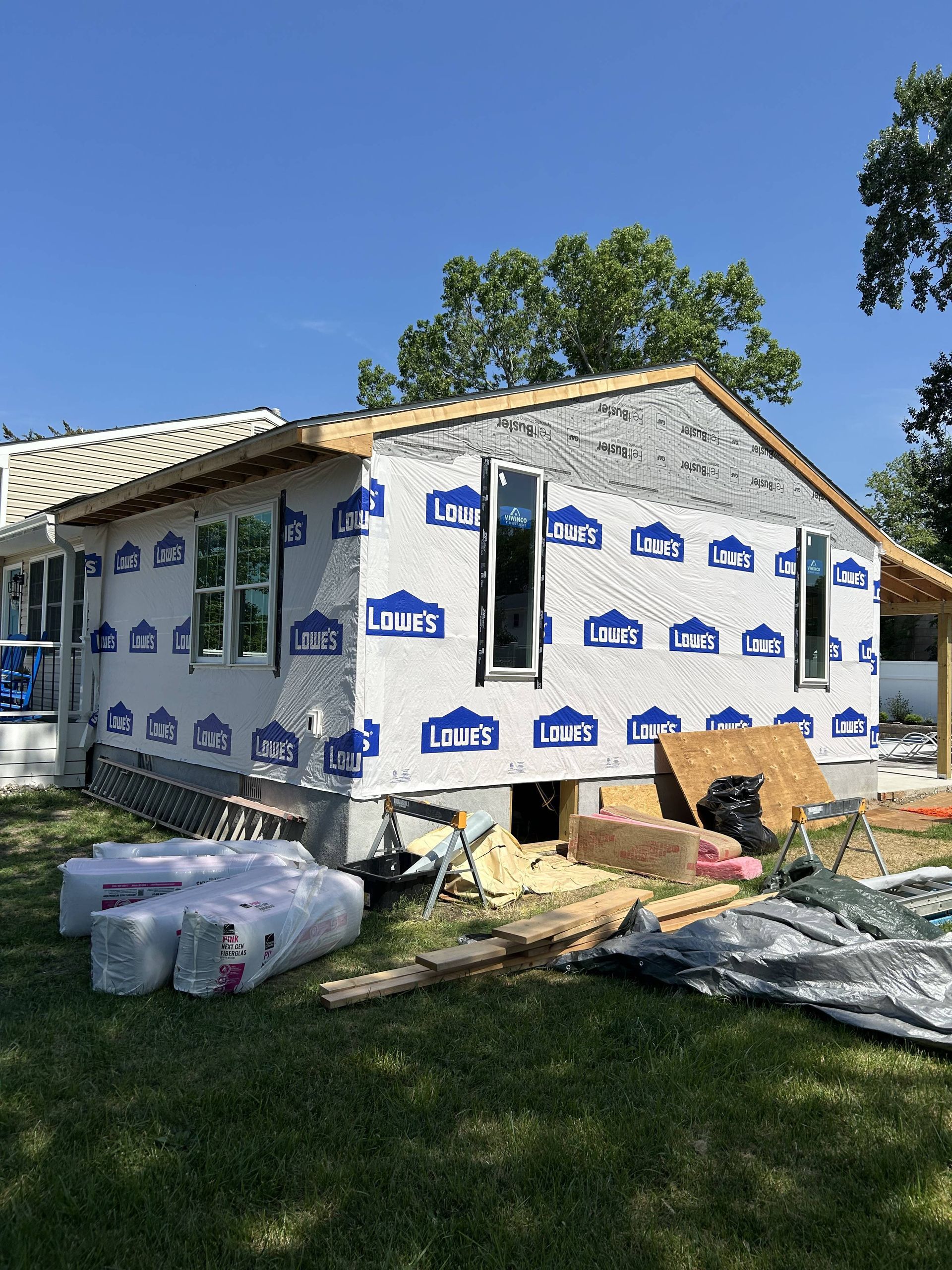 House under construction wrapped in white Tyvek, with roofing materials and debris in the yard.