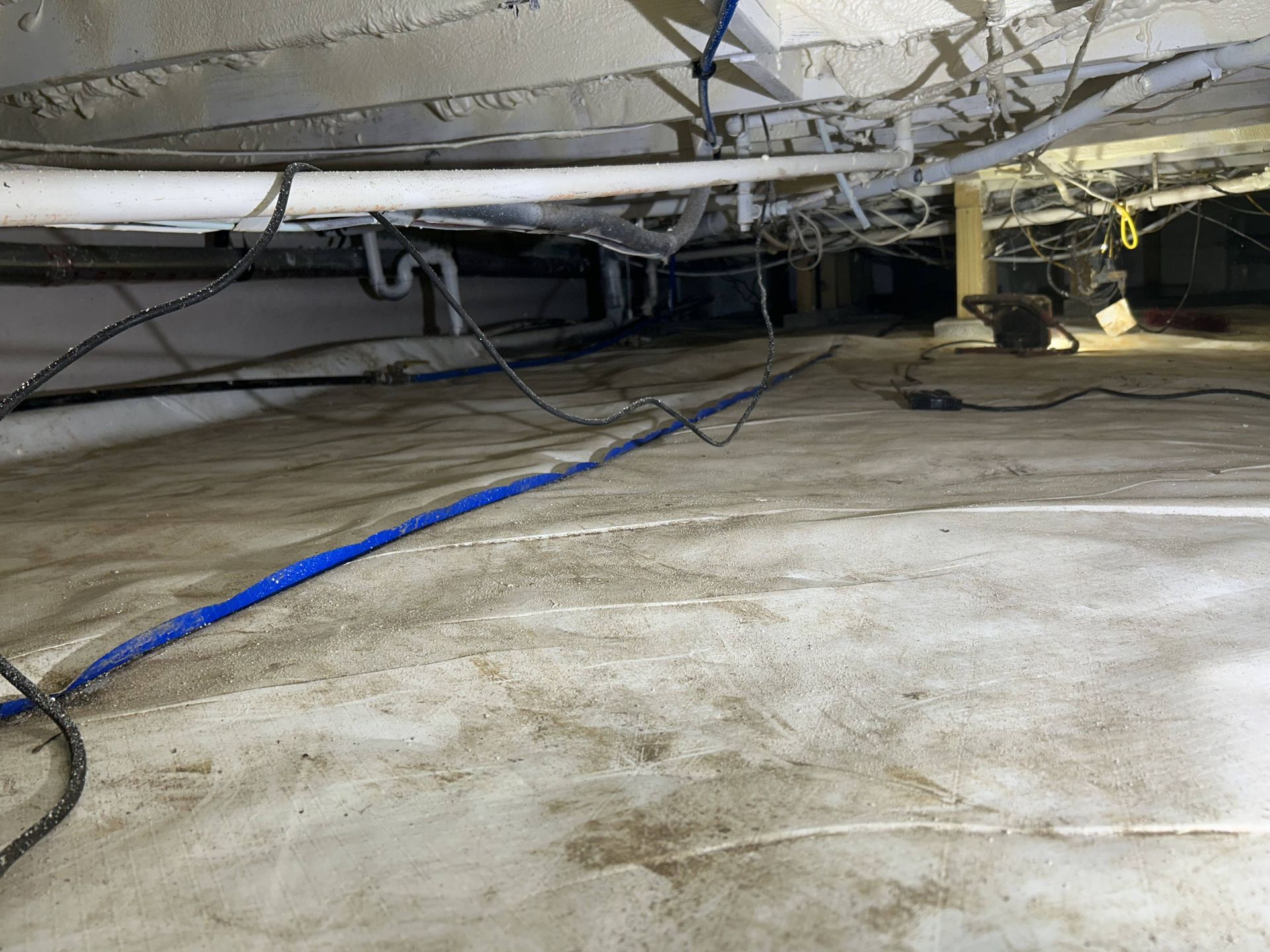 Crawlspace under a house with exposed insulation, wires, and a blue hose or cable across the dirt floor