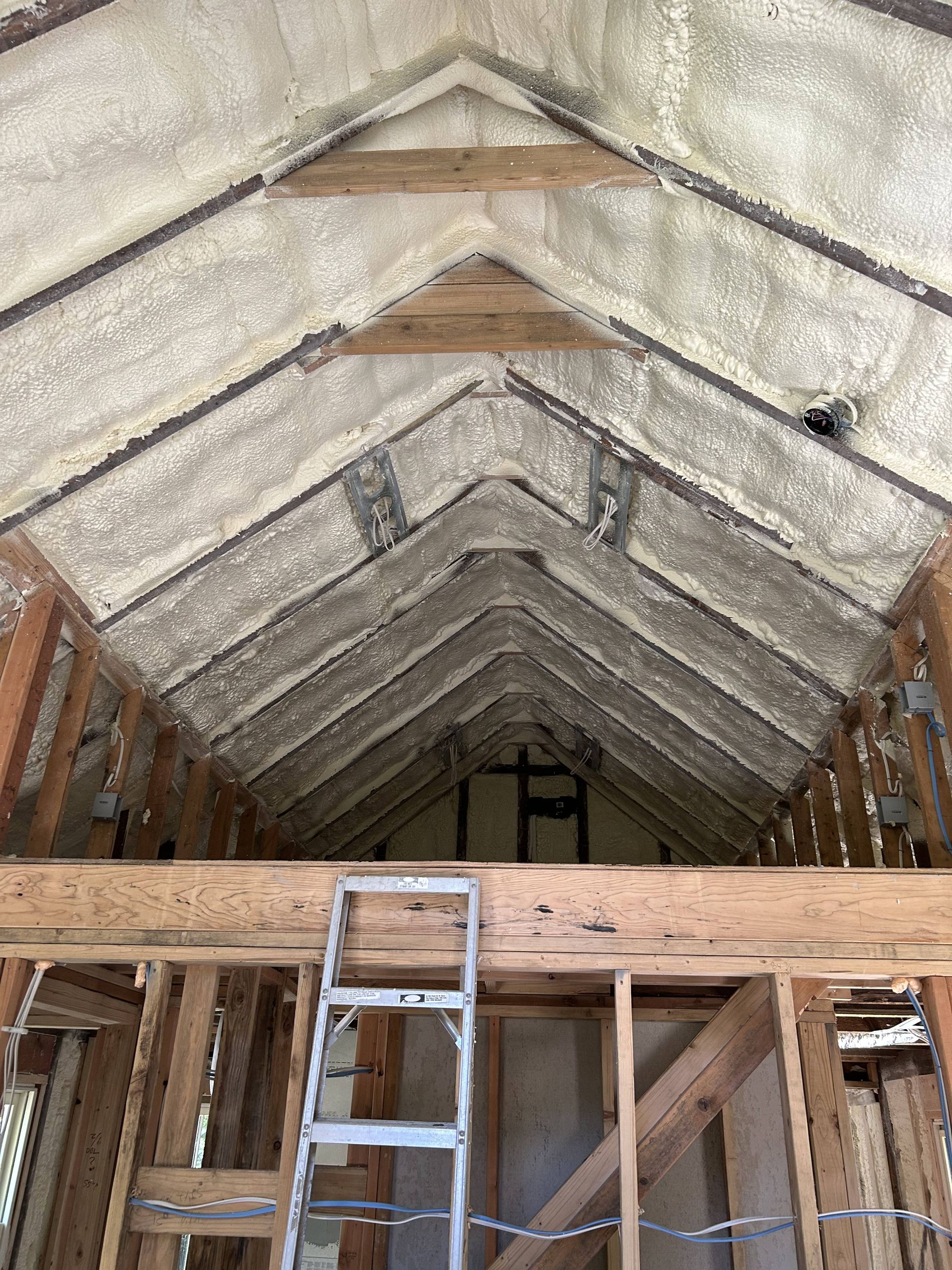 Interior of a house under construction with exposed roof trusses, insulation, and a ladder on the floor.