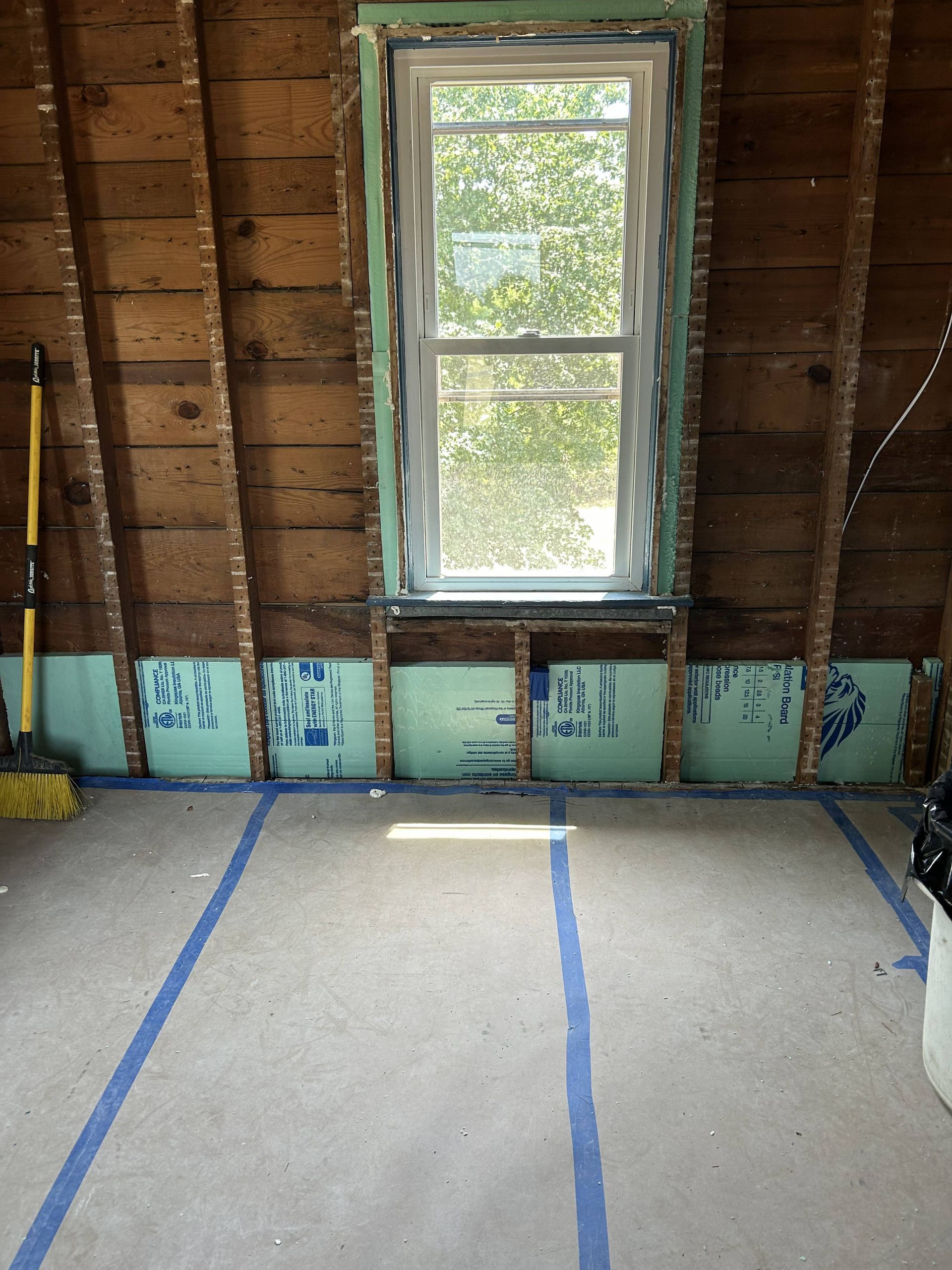 Unfinished room with exposed walls, blue tape on floor, and a window letting in daylight.
