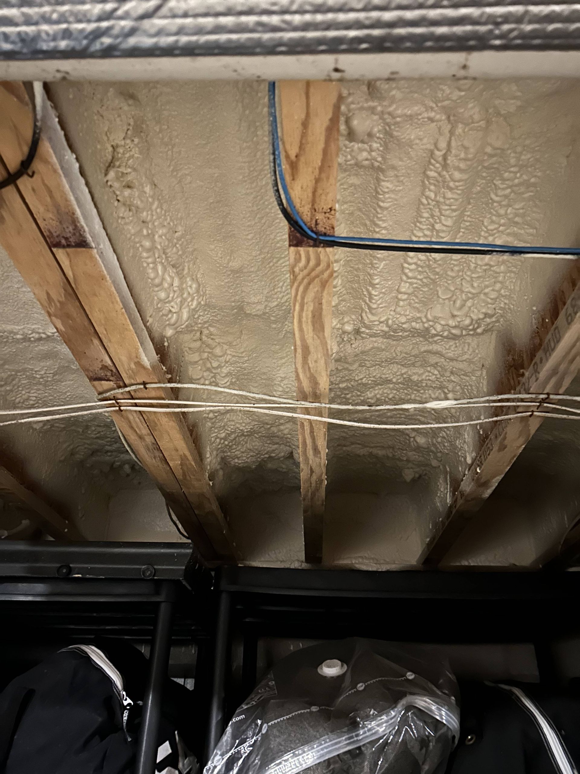 Basement ceiling with exposed spray foam insulation, wooden joists, and hanging cables above storage shelves