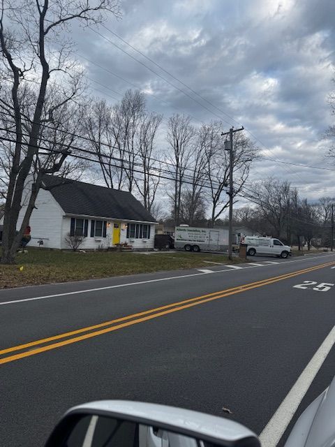 Rural roadside with white house, parked trucks, leafless trees, and a cloudy sky.