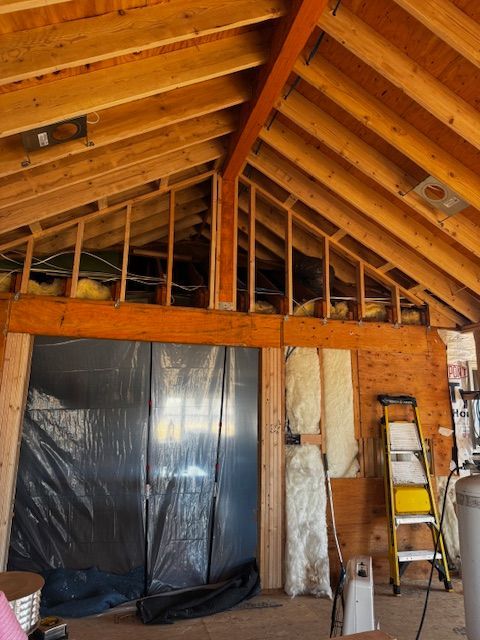 Interior of a wooden attic under renovation, with exposed rafters, insulation, plastic sheeting, and a ladder.