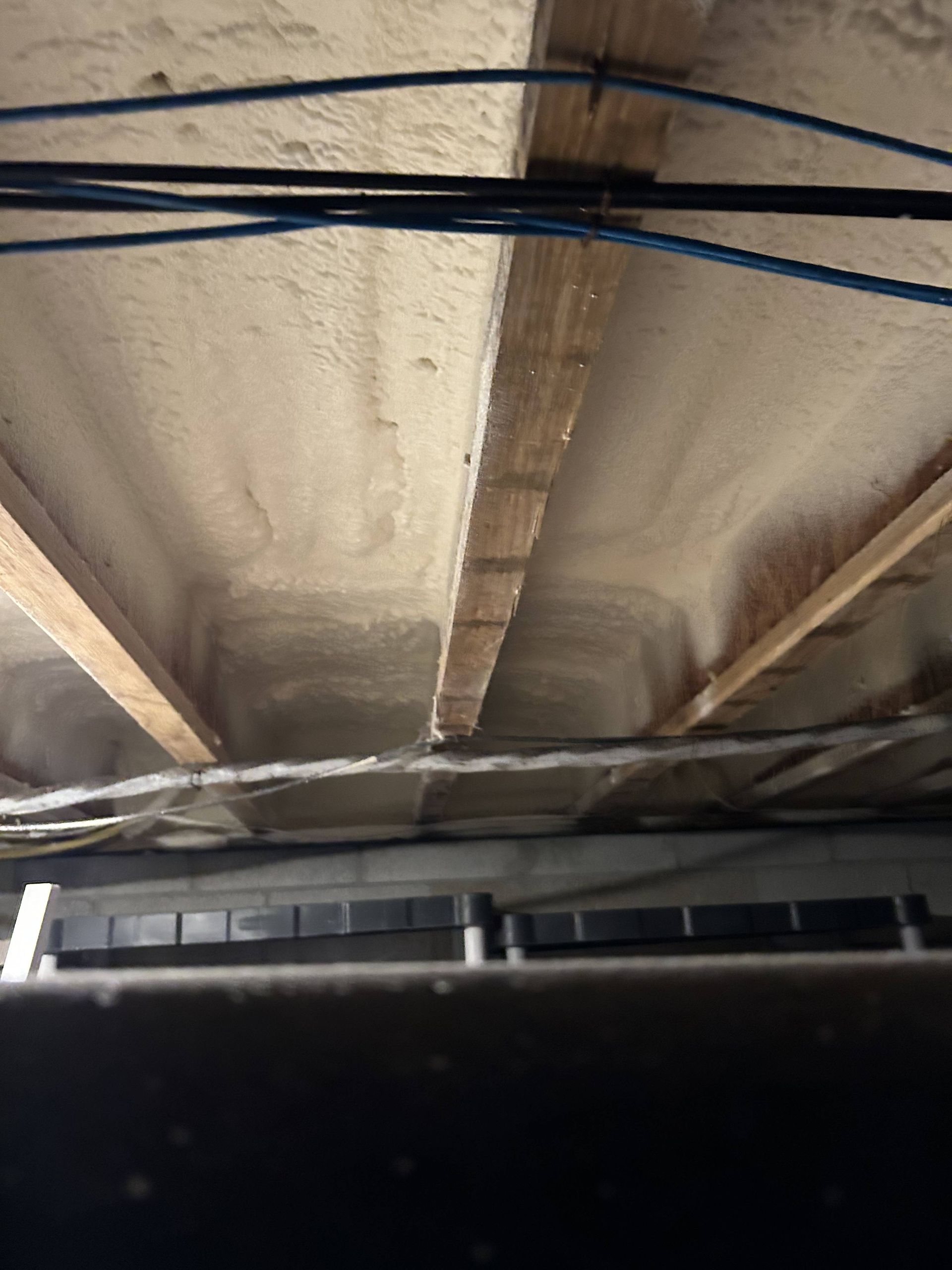 Underside of a roof with wooden beams and spray insulation, with dark cables running across it