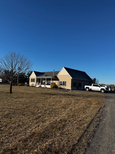 Single-story tan house with a dark roof beside a dirt driveway under a clear blue sky