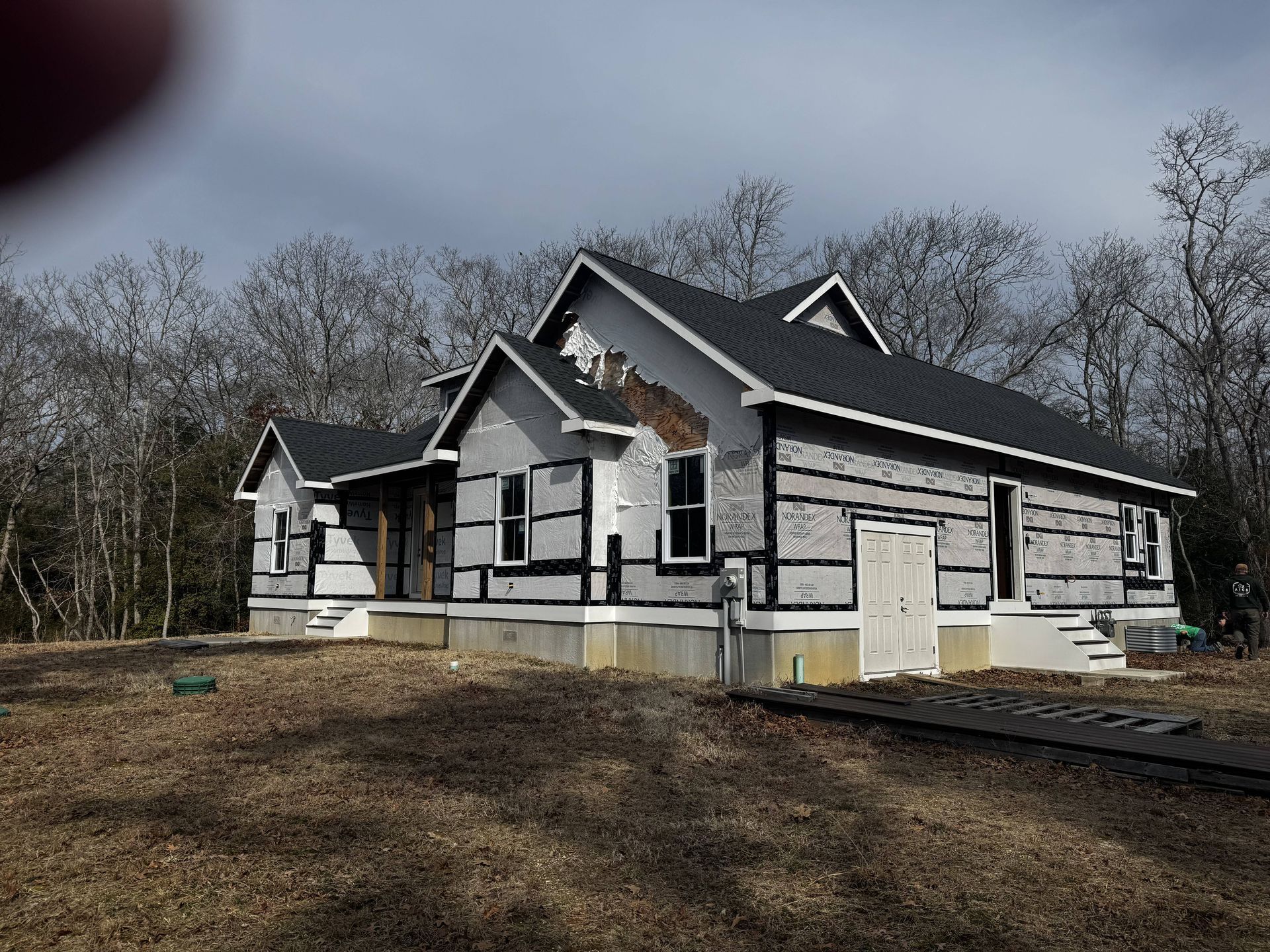 Partially finished house with gray siding and dark roof on a bare lot under cloudy sky
