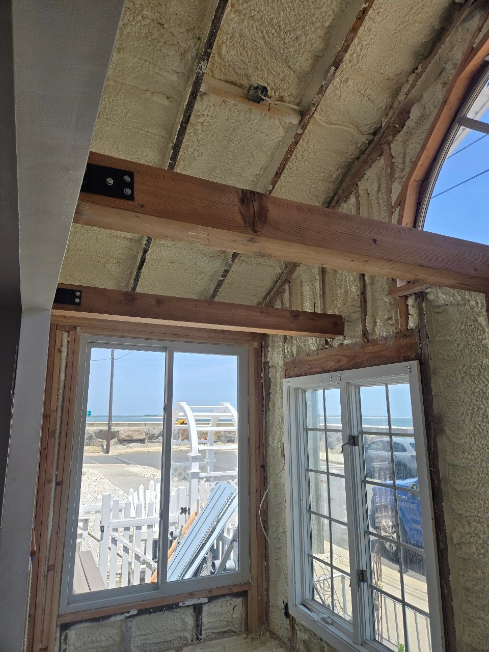 Interior of a narrow room with exposed insulation, wooden beams, and windows overlooking a bright waterfront scene