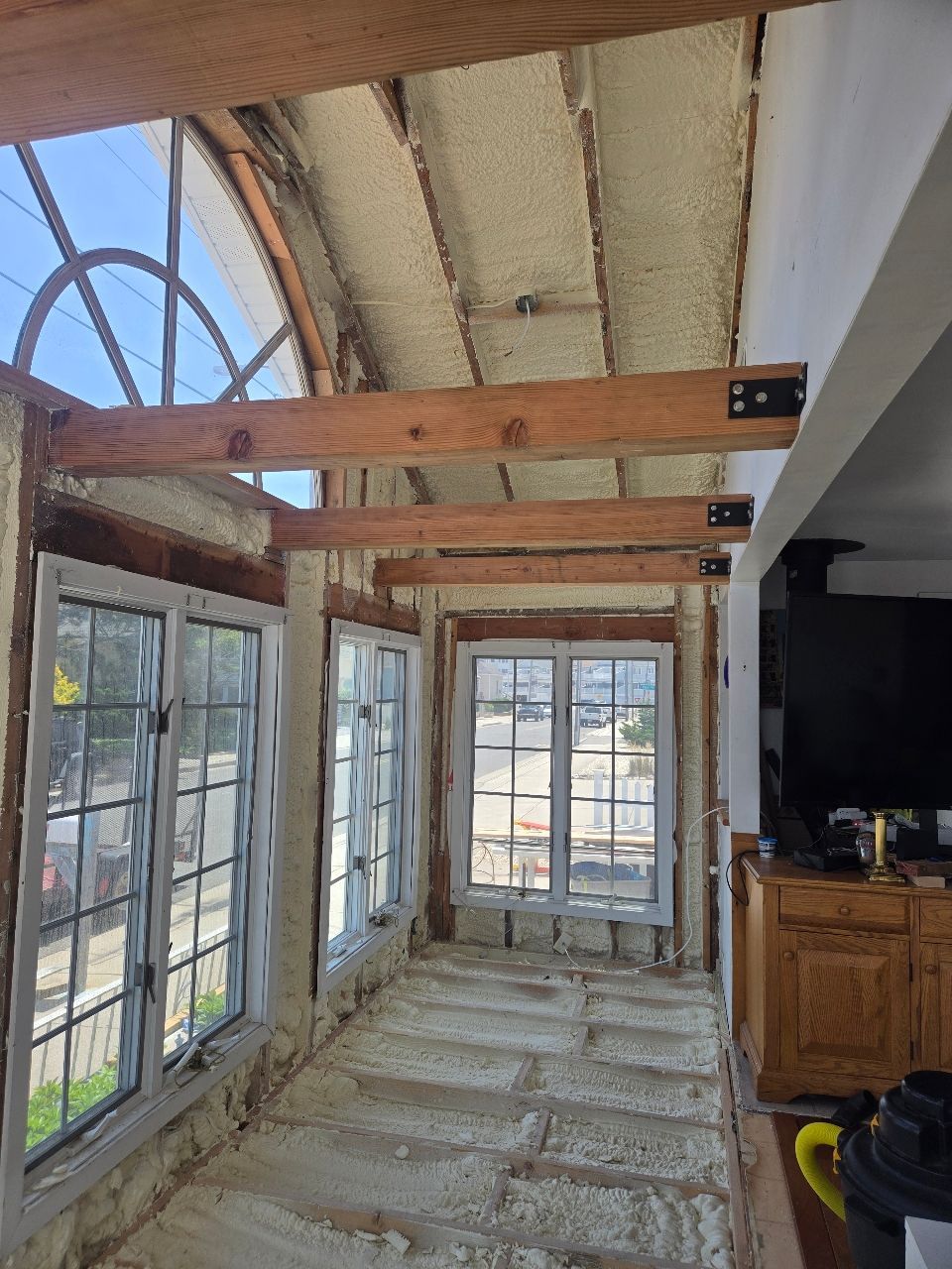 Sunroom under renovation with exposed beams, unfinished floor, and tall windows overlooking outdoors