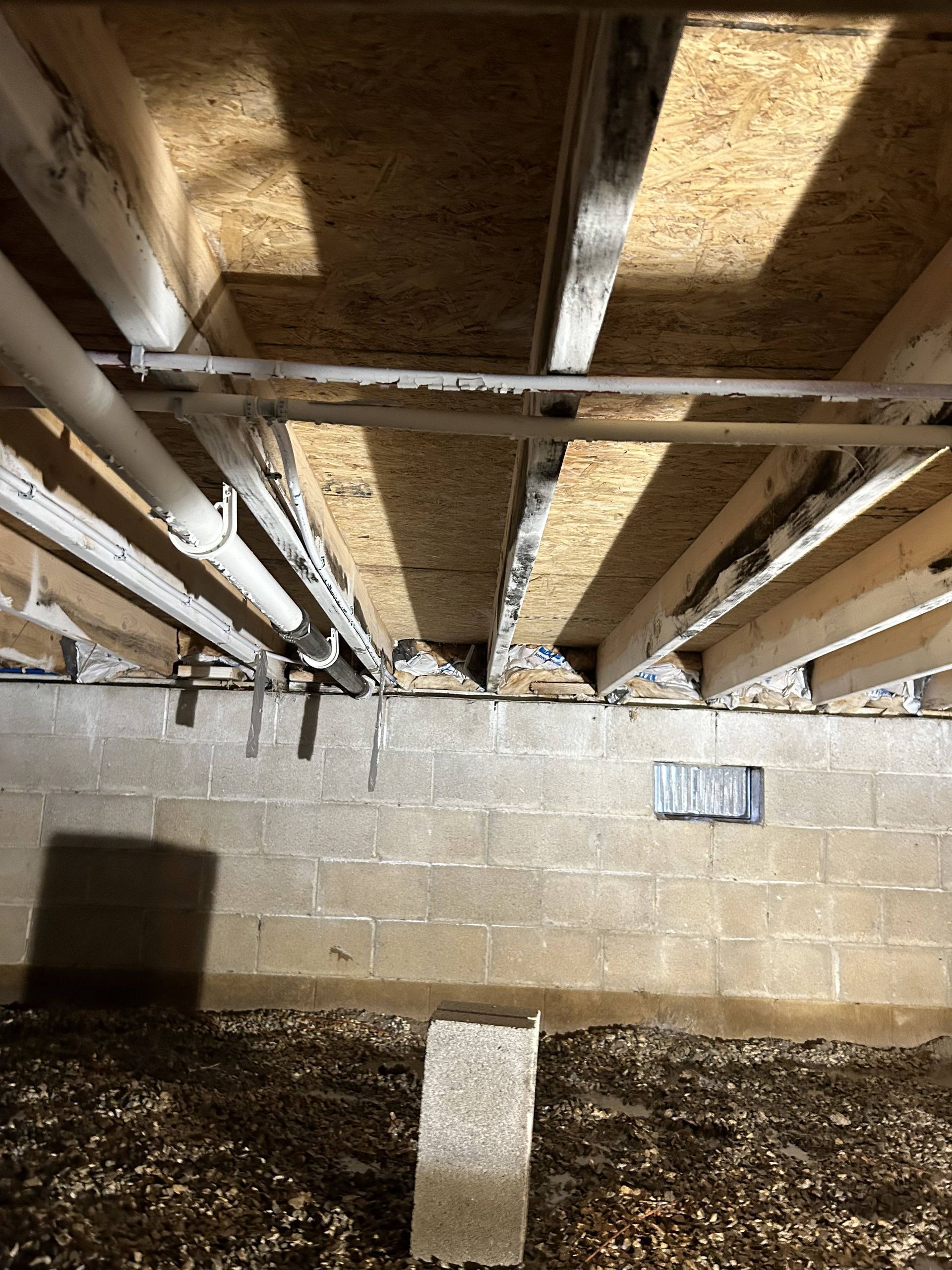Basement crawl space with exposed wooden joists, pipes, and cinderblock wall under a floor opening