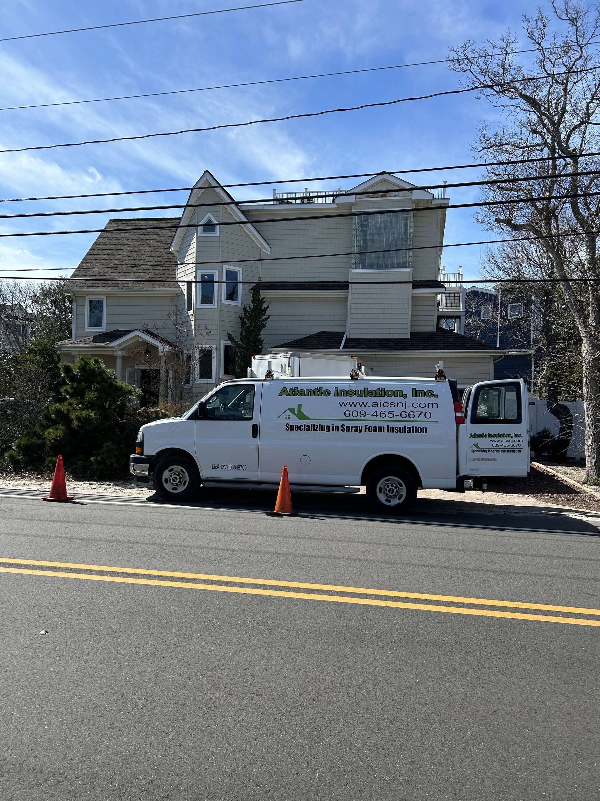 White utility van parked by a large house, with orange cones and a tree-lined street under clear skies.