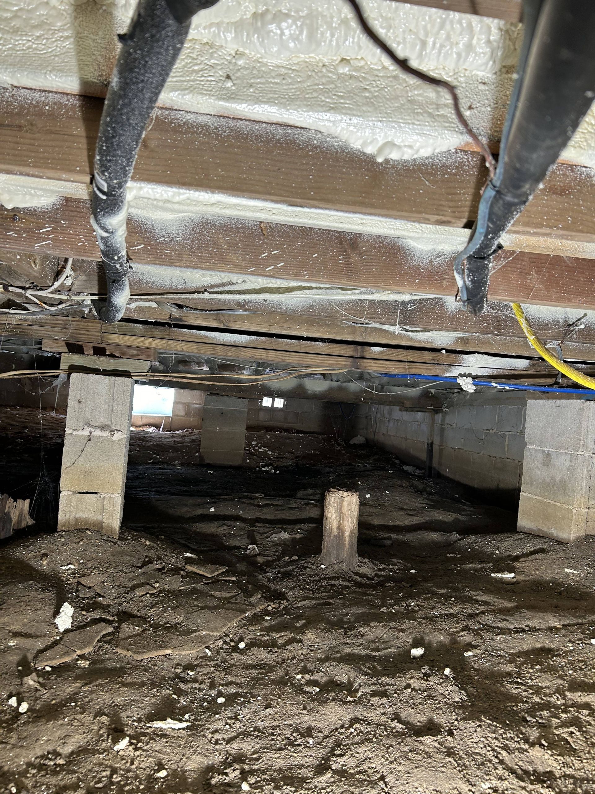 Crawlspace under a floor with exposed beams, support posts, dirt ground, and some wiring.