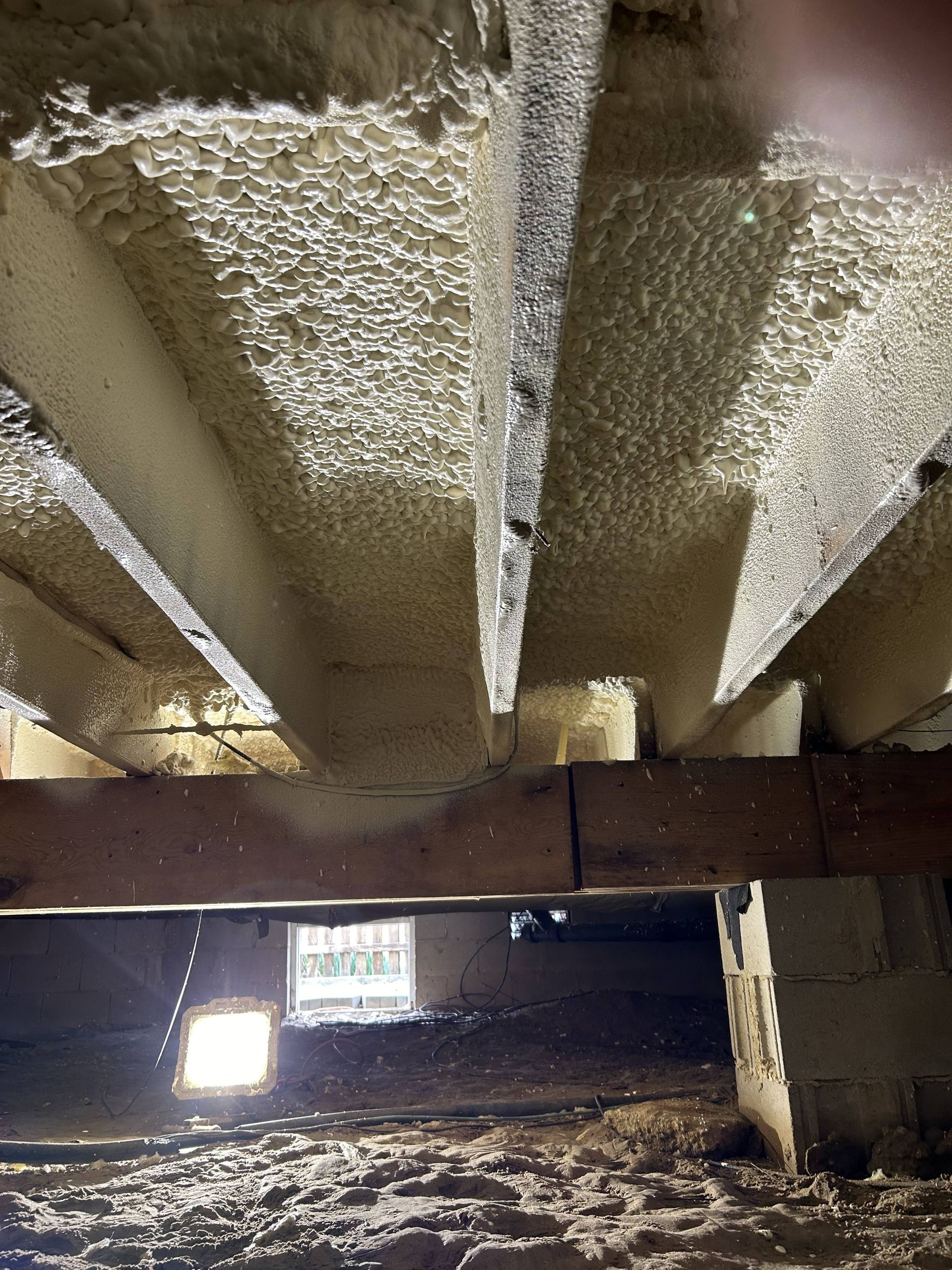 Insulated crawl space with exposed wooden beams and dirt floor, lit by a small window.