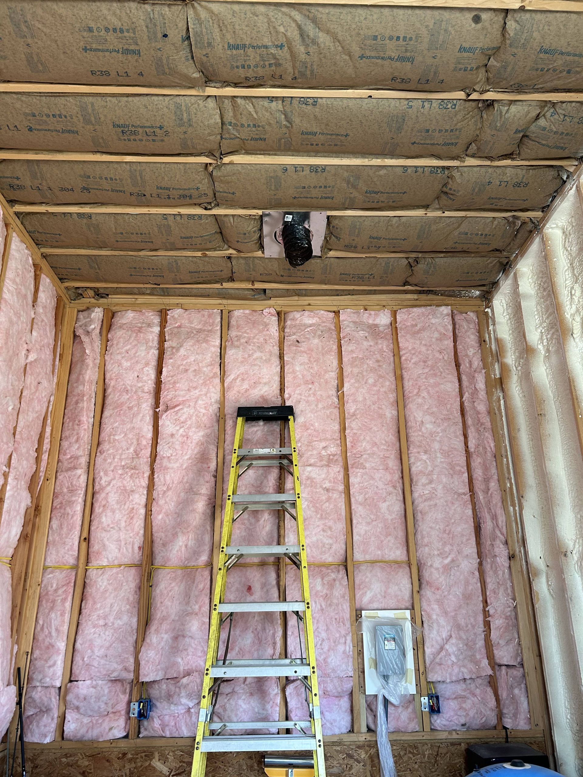 Pink-insulated attic with a ladder below a small roof opening and unfinished wood ceiling.