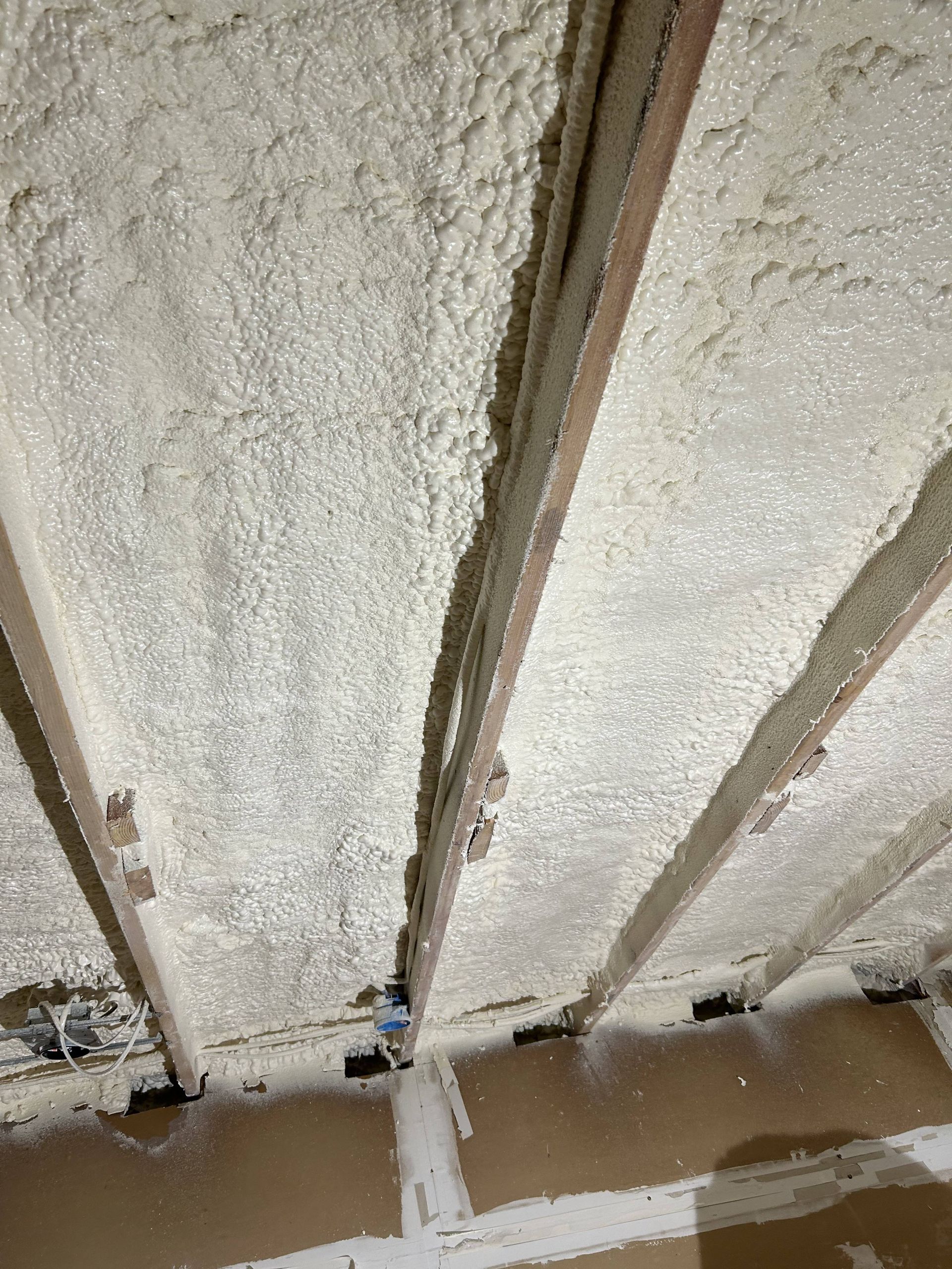 Insulated wall framing with white foam spray insulation between vertical wooden studs and a concrete floor edge