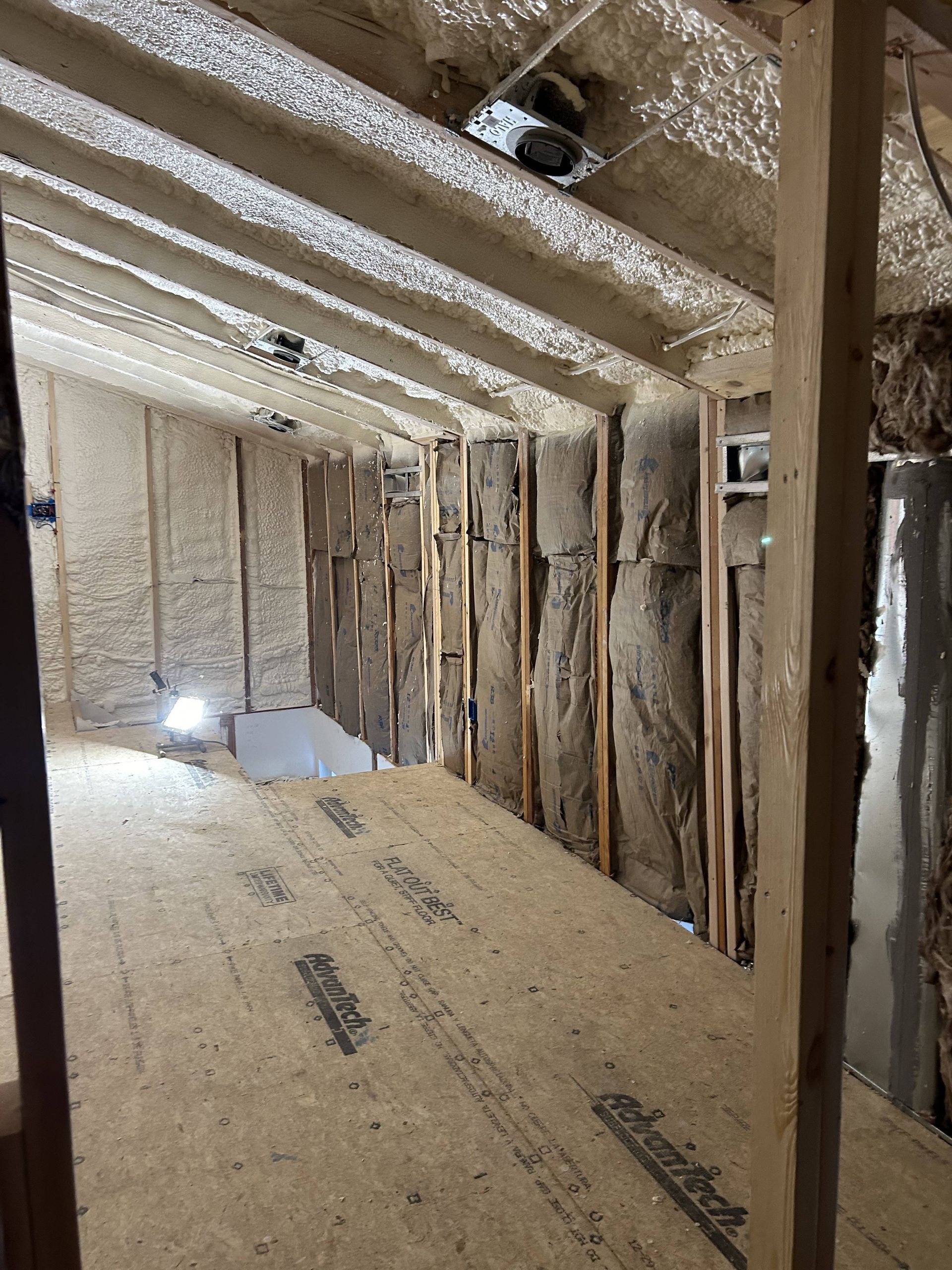 Damaged unfinished attic with exposed insulation, roof beams, and plywood floor, lit by a small light.