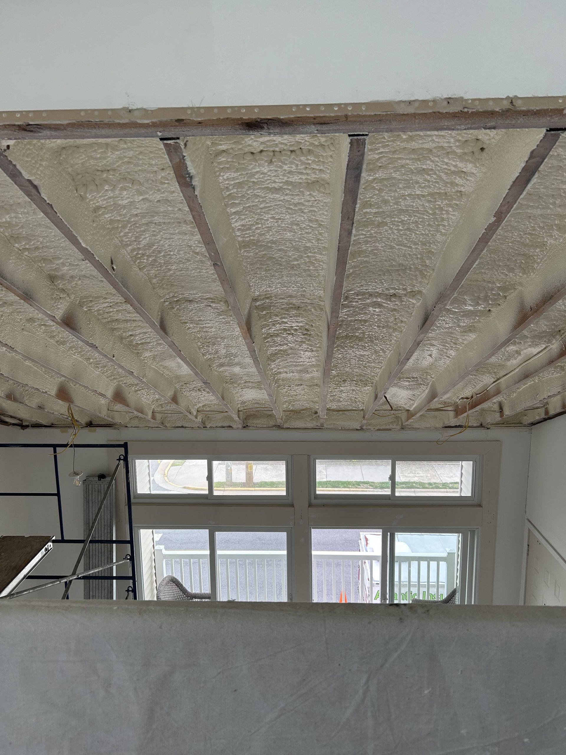 Insulated ceiling under construction above a room with windows and a ladder, partially covered by a wall.