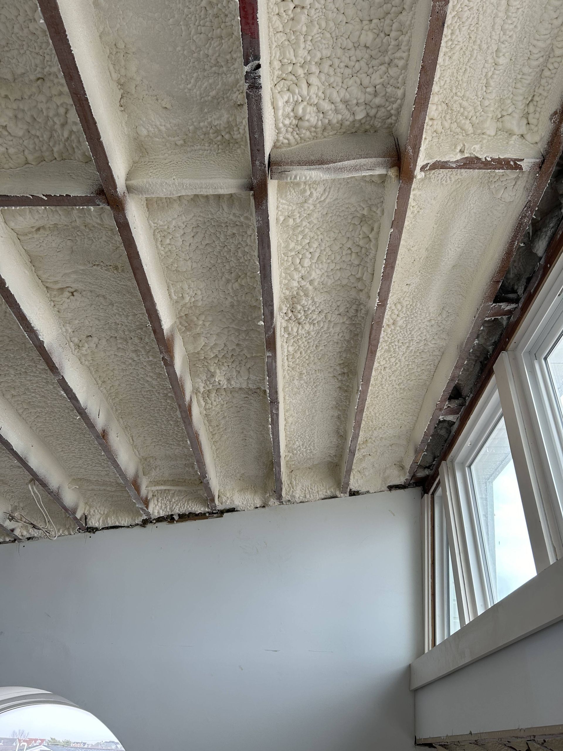Unfinished insulated ceiling with exposed beams above a room with white walls and a window.