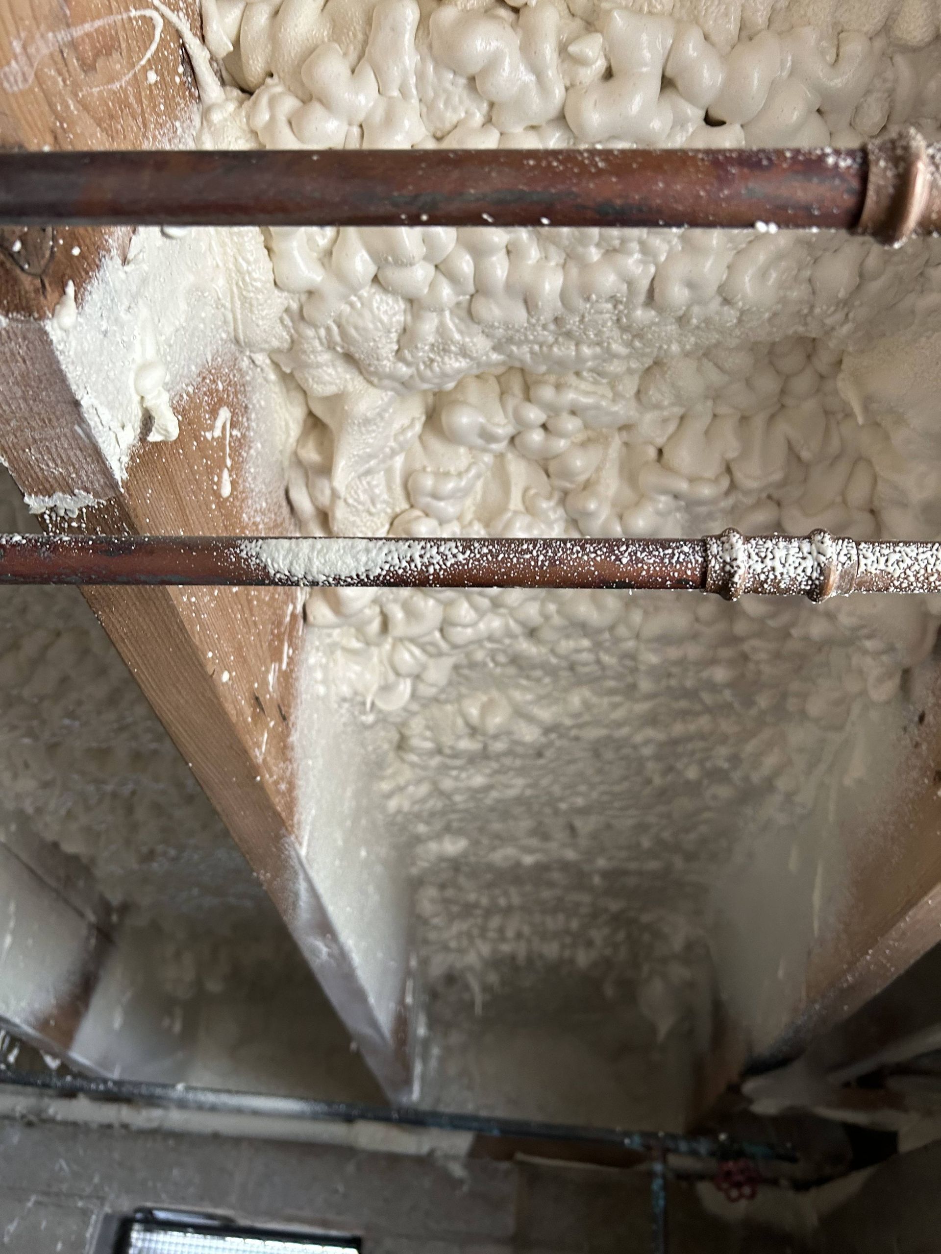 Foam insulation on attic ceiling with exposed brown pipes and rafters