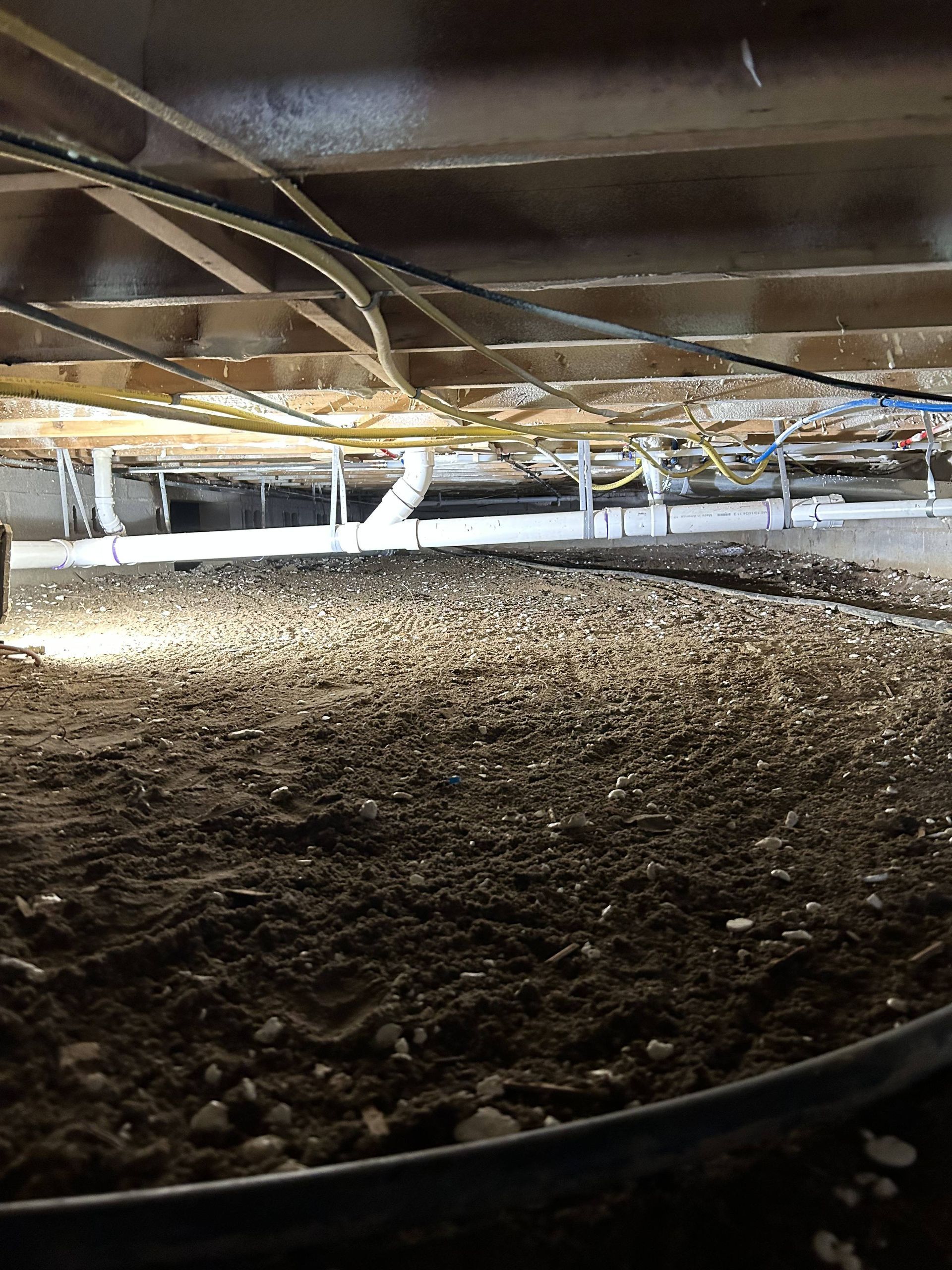 Low crawl space with dirt floor, exposed wooden beams, and pipes overhead