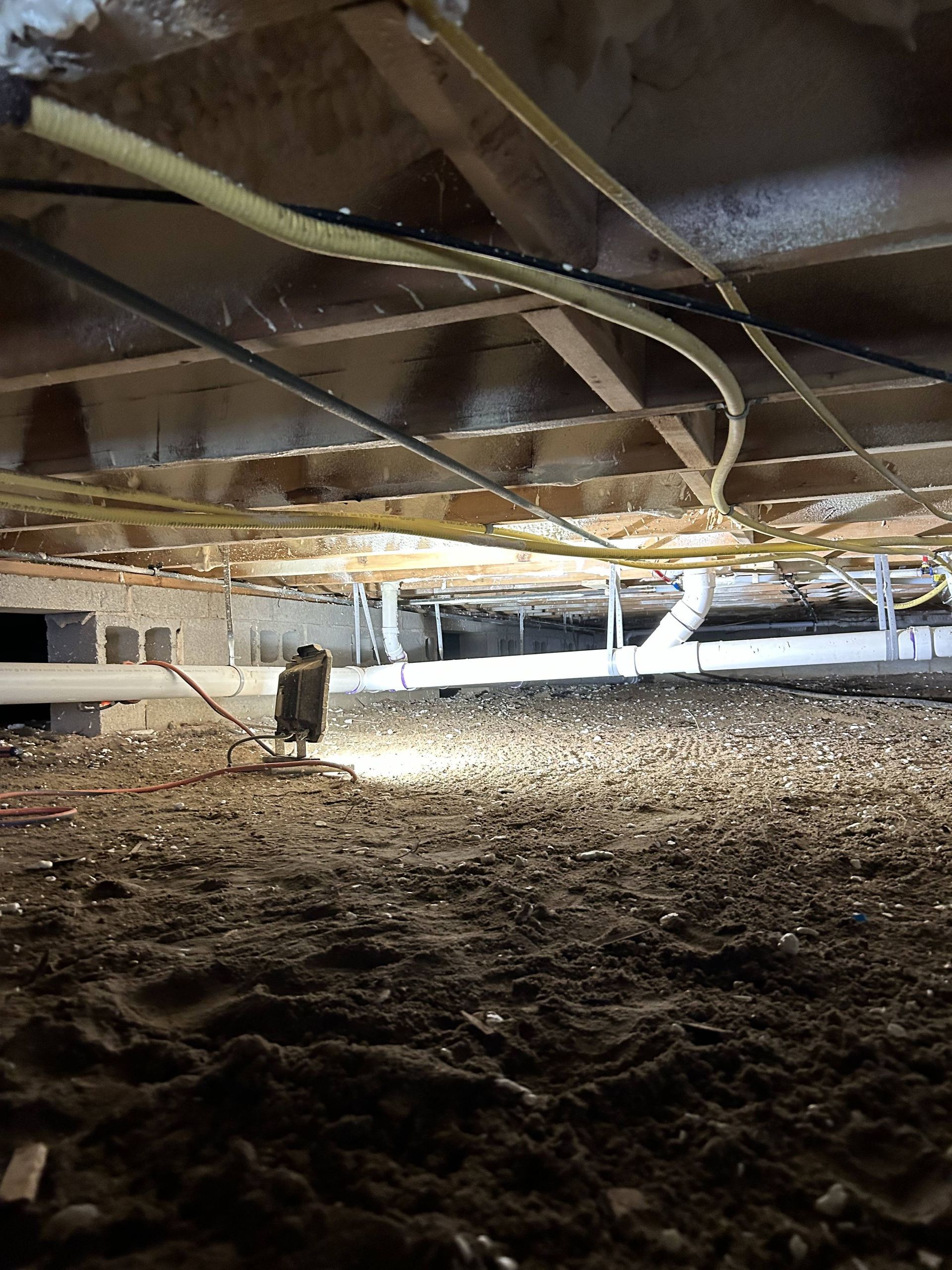 Crawlspace under a house with exposed wooden beams, ducts, and dirt floor illuminated by a flashlight