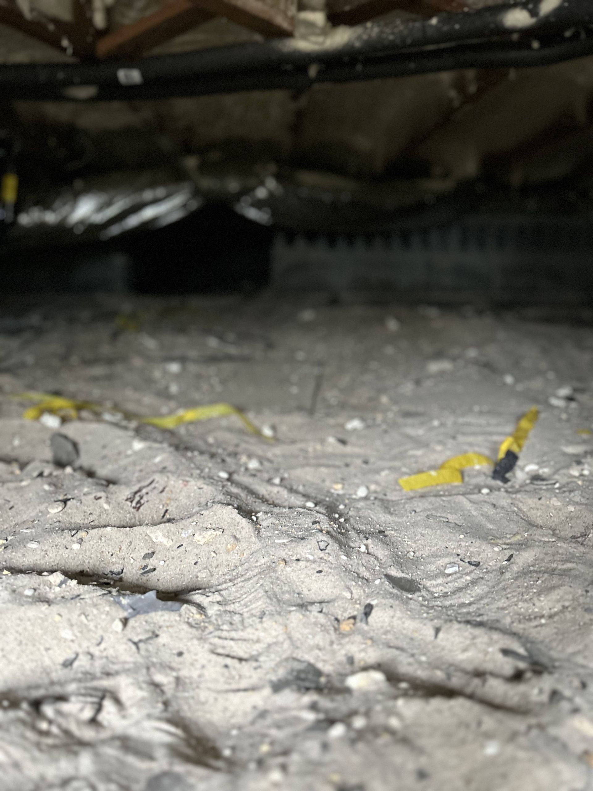 Close-up of dusty debris under a dark structure, with small yellow fragments scattered on the floor.